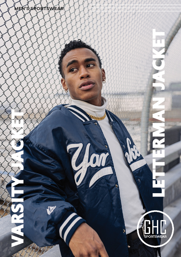 A young man wearing a stylish custom men's varsity jacket, also known as a letterman jacket, in a navy blue color with white detailing, standing against a chain-link fence, showcased on ghcsportswear.com.