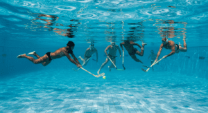 Underwater hockey being played at the bottom of a swimming pool, with players using sticks to push a puck across the floor, no breathing gear allowed."